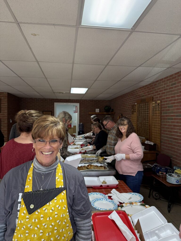 Smiling volunteer in a yellow apron at the serving line in the Fellowship Hall