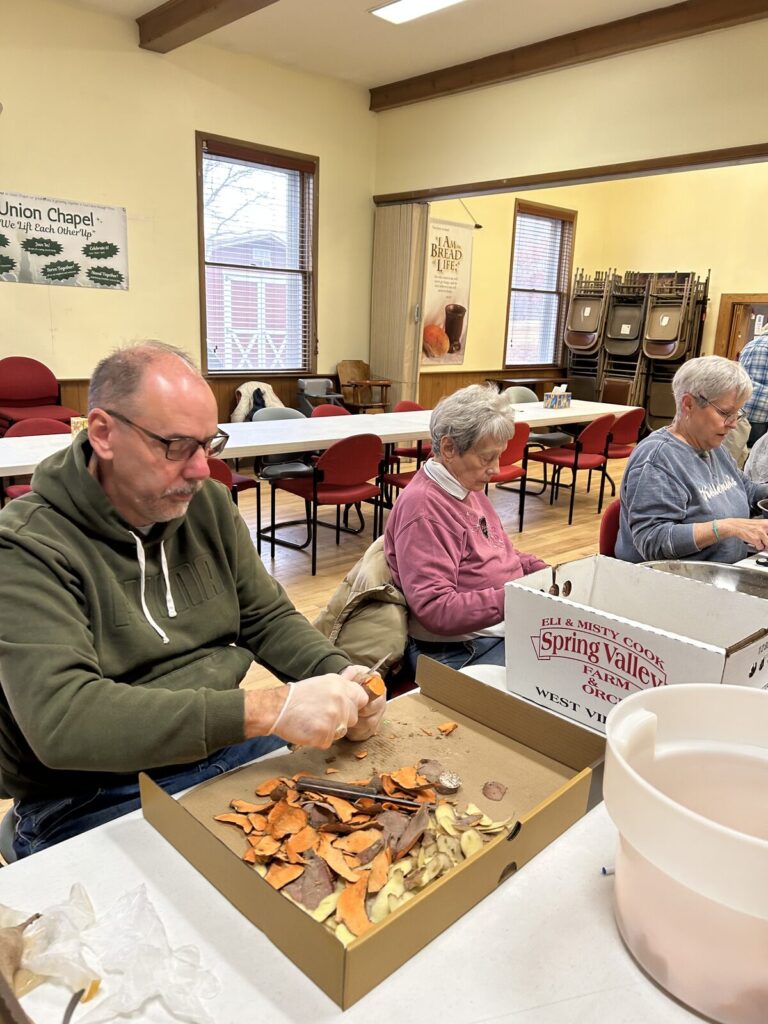 Volunteers seated at tables preparing sweet potatoes in the Fellowship Hall