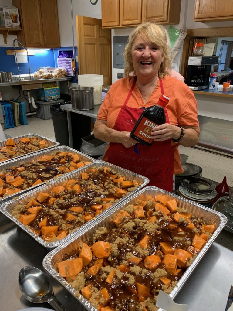 Volunteer in a red apron behind large pans of baked sweet potatoes