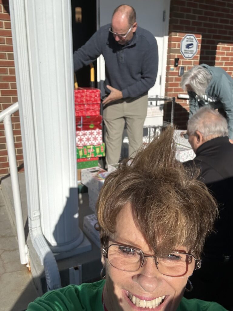Smiling volunteer takes a selfie outside the church entrance while other volunteers load stacks of Operation Christmas Child shoeboxes